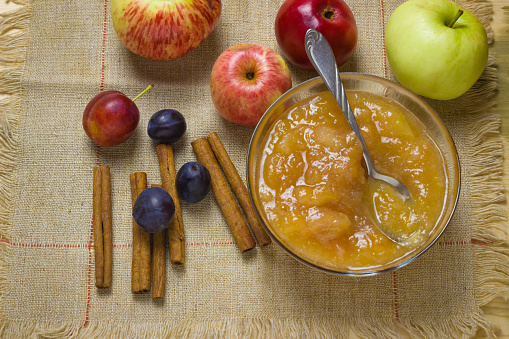 Homemade apple jam. Traditional jam in a glass bowl. Napkin made of harshness. Ripe apples and plums, cinnamon sticks. Wood background.