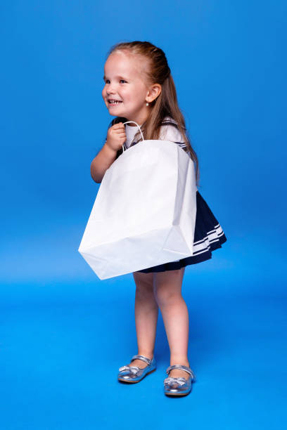 Smiling girl with long hair, wearing a dress, looking in a white shopping bag, on a blue background. stock photo