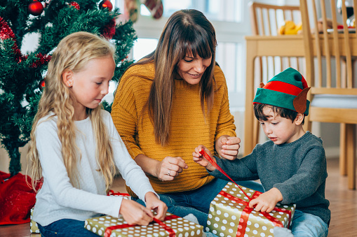 Two cute children and their beautiful mother sitting on the floor near the Christmas tree and opening gifts.