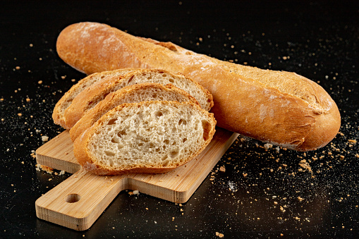 Pieces of fresh baguette on a wooden board. Delicious bread prepared to make sandwiches. Dark background.