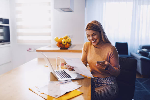 Cute caucasian smiling blonde woman in sweater holding bills in one hand and in other smart phone. On table are laptop and bills. Apartment interior. Cute caucasian smiling blonde woman in sweater holding bills in one hand and in other smart phone. On table are laptop and bills. Apartment interior. house phone stock pictures, royalty-free photos & images