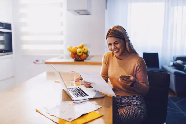 Photo of Cute caucasian smiling blonde woman in sweater holding bills in one hand and in other smart phone. On table are laptop and bills. Apartment interior.