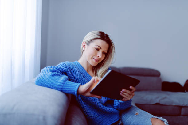 Beautiful caucasian smiling blonde in blue sweater and jeans sitting on sofa in living room and using tablet. Apartment interior. Beautiful caucasian smiling blonde in blue sweater and jeans sitting on sofa in living room and using tablet. Apartment interior. graphics tablet stock pictures, royalty-free photos & images