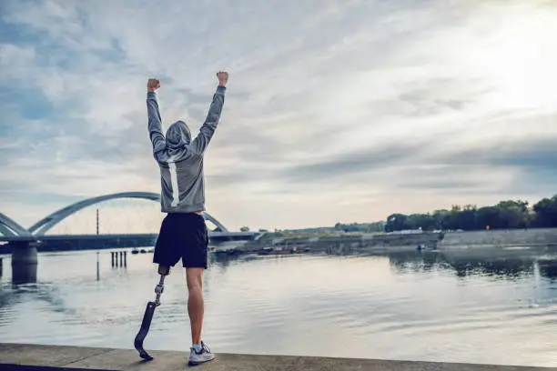 Highly motivated caucasian handicapped sportsman in sportswear and with artificial leg standing on quay and holding arms up. Only way to win is to be yourself.