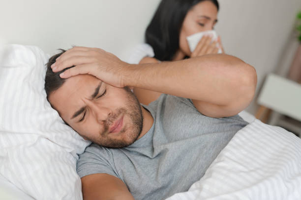 giovane coppia che ha l'influenza e giace a letto a casa. l'uomo ha mal di testa mentre sua moglie starnutisce al tovagliolo - cold and flu flu virus bed couple foto e immagini stock