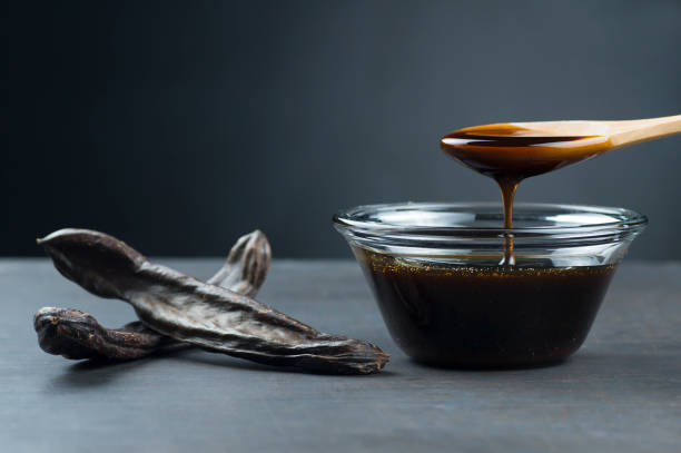 carob molasses in glass bowl and in wooden spoon and carob pods on rustic background, locust bean healthy food, ceratonia siliqua ( harnup ) - ceratonia imagens e fotografias de stock