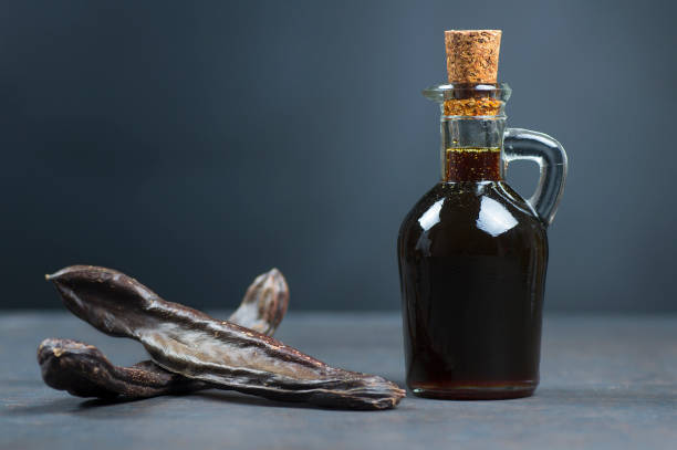 glass bottle of carob molasses and carob pods on rustic background, locust bean healthy food, ceratonia siliqua ( harnup ) - ceratonia imagens e fotografias de stock