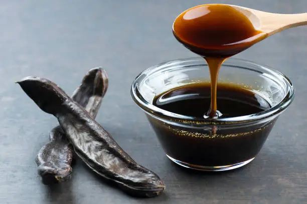 Photo of carob molasses in glass bowl and in wooden spoon and carob pods on rustic background, locust bean healthy food, Ceratonia siliqua ( harnup )