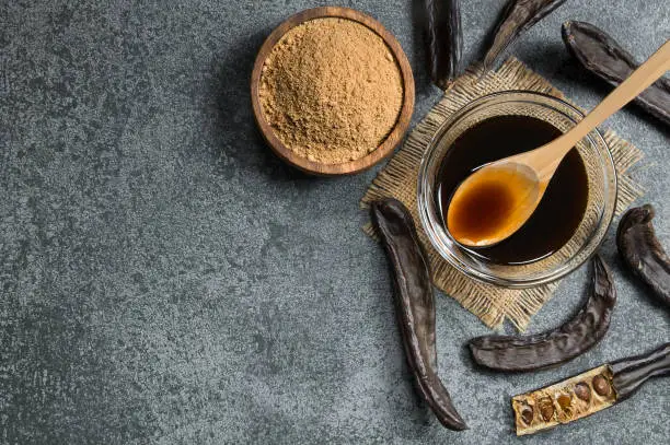 Photo of carob molasses in glass bowl and carob powder or flour with carob pods on rustic background, locust bean healthy food, Ceratonia siliqua ( harnup )