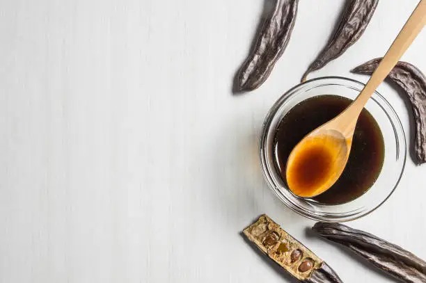 Photo of carob molasses in glass bowl and in wooden spoon and carob pods on rustic background, locust bean healthy food, Ceratonia siliqua ( harnup )