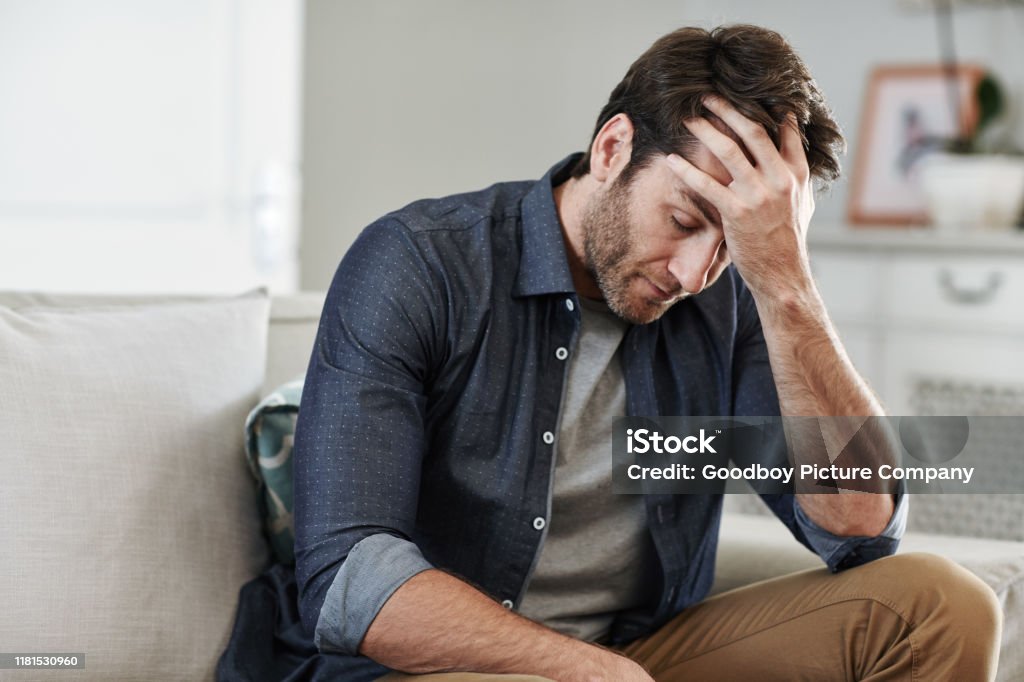 Man sitting alone at home looking sad and distraught Man looking depressed while sitting alone with his head in his hand on his living room sofa at home Men Stock Photo