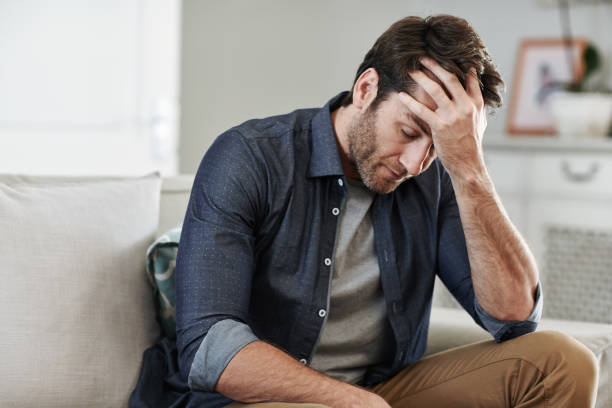 Man sitting alone at home looking sad and distraught Man looking depressed while sitting alone with his head in his hand on his living room sofa at home grim stock pictures, royalty-free photos & images