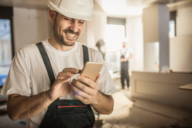 Happy construction worker using cell phone during home renovation. Happy male worker text messaging on smart phone while being in renovating home. craftsperson stock pictures, royalty-free photos & images