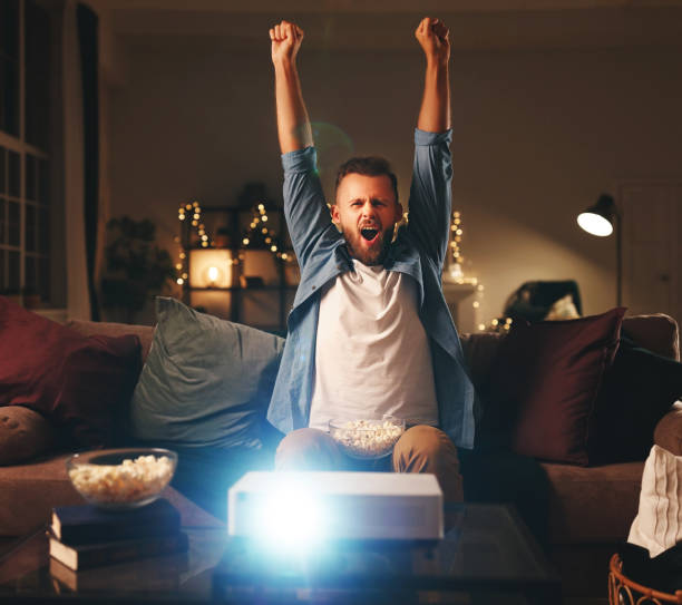 young male cheerleader watching football on tv projector at home in evening - fan sport football male imagens e fotografias de stock