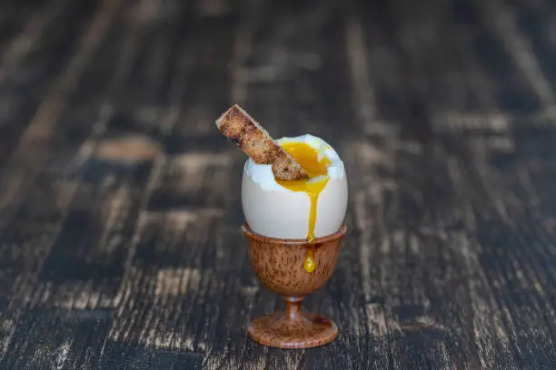 Soft boiled egg in eggcup with slice of toasted bread on wooden table background, close up