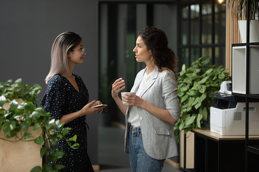 Asian and Caucasian ethnicity businesswoman talking discuss common project standing in workplace hall more experienced employee share knowledge with intern during coffee break in modern office hallway