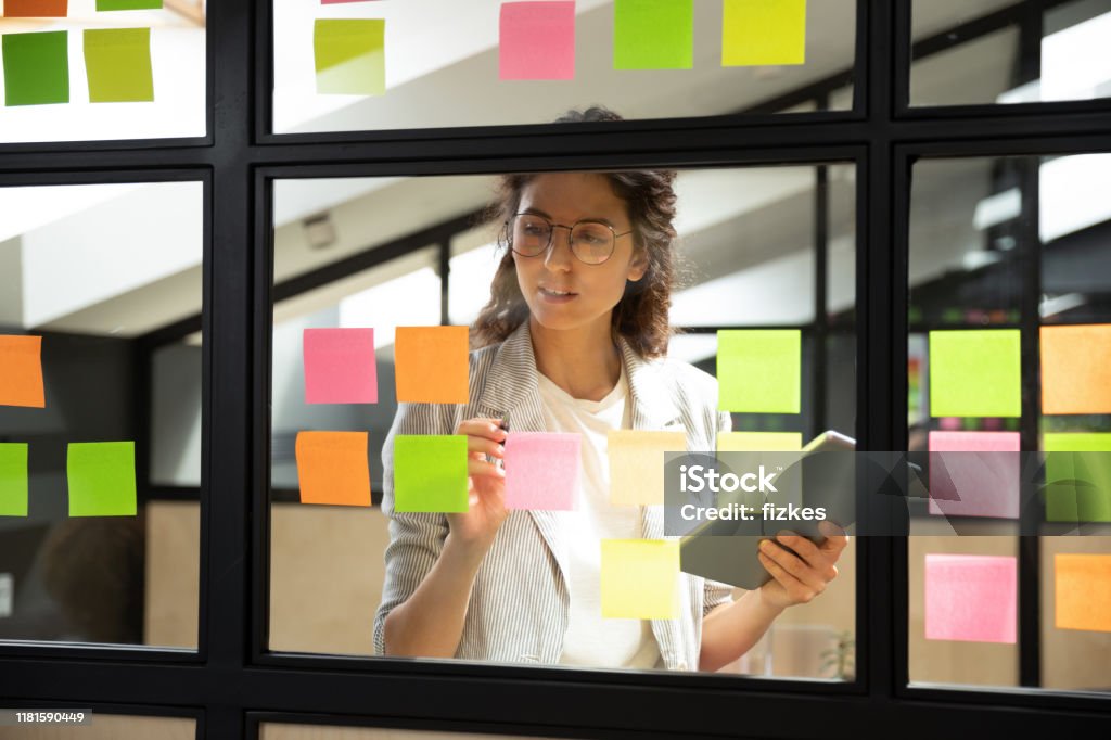 Businesswoman creates priority to-do list on sticky notes Businesswoman creates priority to-do list standing behind glass wall writes fresh ideas interesting creative thoughts on multicolored post-it sticky notes using tablet having fruitful workday concept Chores Stock Photo
