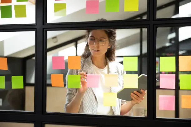 Businesswoman creates priority to-do list standing behind glass wall writes fresh ideas interesting creative thoughts on multicolored post-it sticky notes using tablet having fruitful workday concept