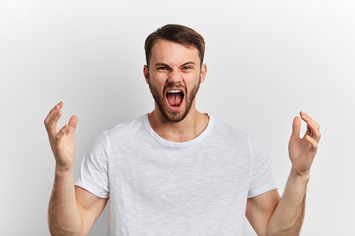 Young man wearing white T-shirt raising his arms, shouting, expressing aggressive expression standing over white isolated background. Frustration concept.I'll kill you