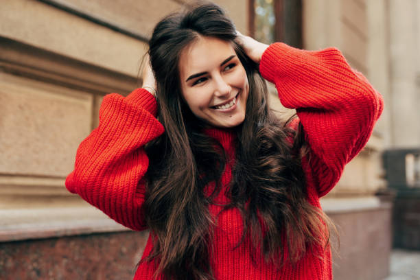 Beautiful young brunette woman smiling broadly with hands on her long hair. Outdoor portrait of pretty female model in trendy knitted red sweater posing during walking in the city street. stock photo