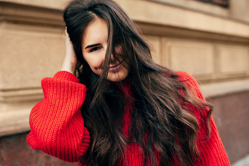 Smiling European brunette woman with long hair looking directly to the camera. Outdoor portrait of blissful female model in trendy knitted red sweater posing during walking in the city street.