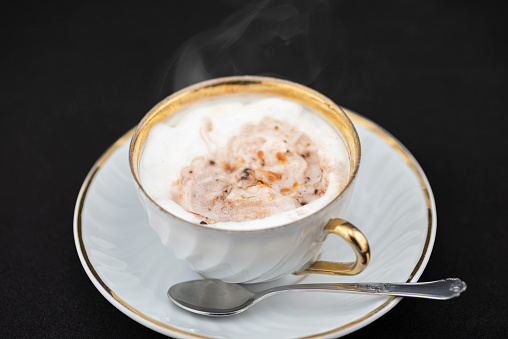 Studio shot of a cappuccino served in an elegant classic style cup with golden borders and a decorated coffee spoon. A pumpkin is represented as latte art. The background is black.