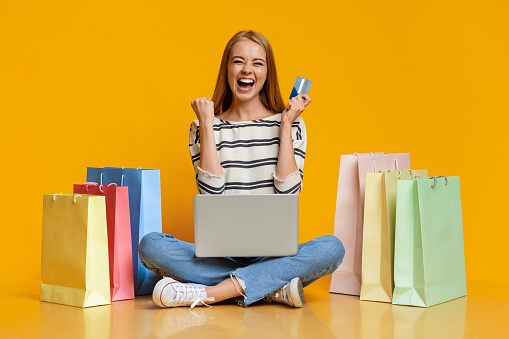 Shopping online. Joyful girl holding credit card and sitting with laptop on laps, surrounded by bright paper bags over orange studio background