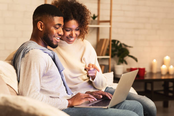 amante afro pareja eligiendo película en el ordenador portátil, descansando en una habitación acogedora - couple home interior laptop computer fotografías e imágenes de stock