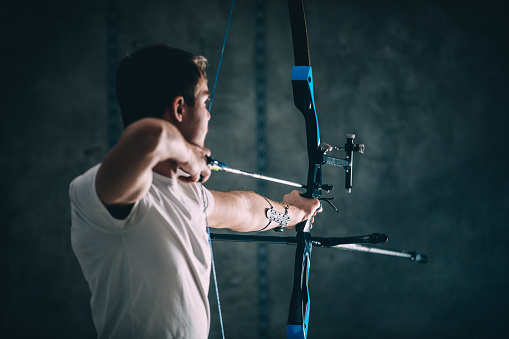 One man, young shirtless archer with bow and arrow training in dark studio alone.