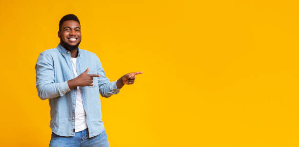 Cheerful black guy pointing two fingers aside at copy space Cool offer. Cheerful african american man pointing two fingers aside at copy space on yellow studio background, panorama men hairstyle afro cool stock pictures, royalty-free photos & images
