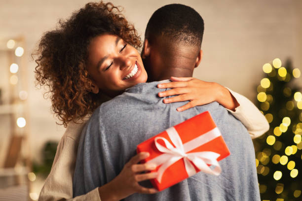 dankbaar afro vrouw met kerstcadeau knuffelen man thuis - christmas people stockfoto's en -beelden