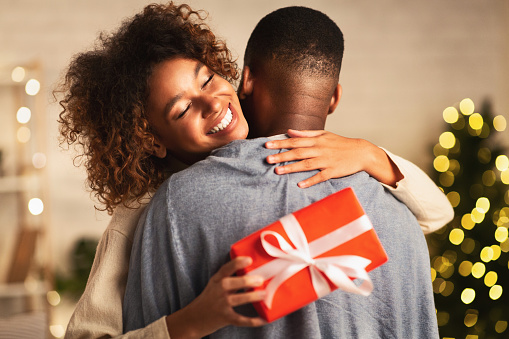 Happy holidays. Thankful afro woman with christmas gift hugging husband at home