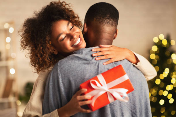 thankful afro woman with christmas gift hugging husband at home - couple black imagens e fotografias de stock