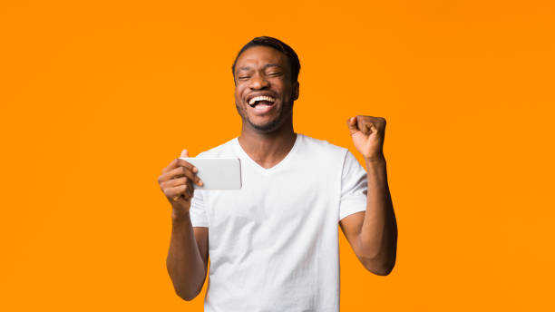 istock Cheerful Black Guy Using Smartphone Shaking Fists, Orange Background, Panorama 1181611148