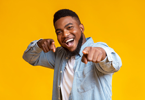 Gotcha. Overjoyed black guy pointing fingers at camera over yellow studio background, empty space