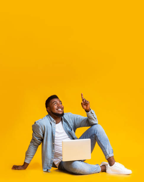 Black guy sitting with laptop and pointing at copy space Great offer. Black millennial guy sitting on floor with laptop and pointing upwards at copy space on yellow background excitement laptop stock pictures, royalty-free photos & images