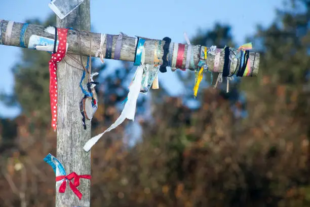 Wooden cross in Camino de Santiago, multicolored ribbons. Bows and tied knots. Copy space on the right. Pine trees and clear sky in the background. Lugo province, Galicia, Spain.