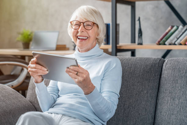 istock Senior woman looking and laughing at her digital tablet on sofa 1181670448