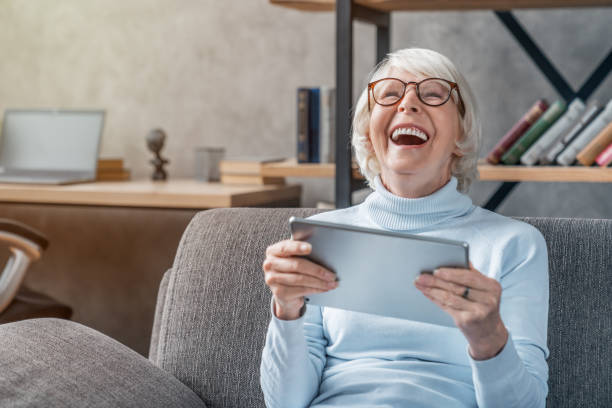 happy senior woman looking and laughing at her digital tablet on sofa - digital tablet women enjoyment happiness imagens e fotografias de stock