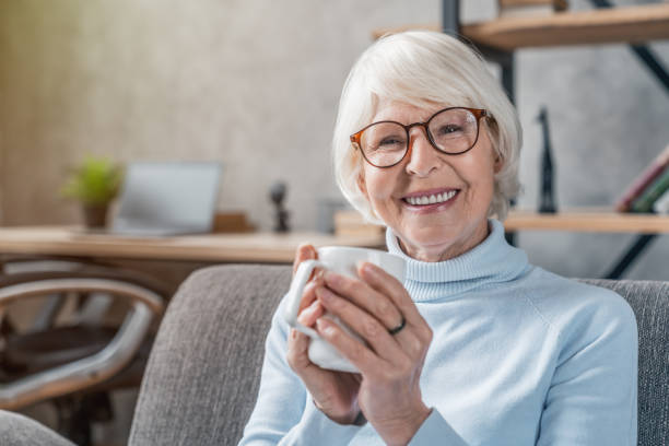 mujer mayor bebiendo bebida caliente sentada en el sofá en casa - senior living communitiy fotografías e imágenes de stock