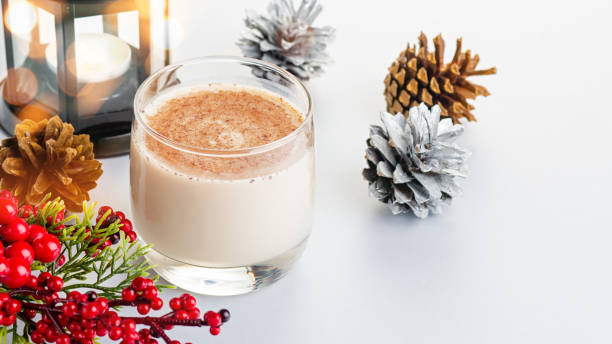 cocktail tradizionale natalizio eggnog con uova, alcol, noce moscata grattugiata e primo piano alla cannella. bevanda tradizionale dolce su tavolo grigio con decorazioni beige e rosse e pigne - luxury milk dinner glass foto e immagini stock