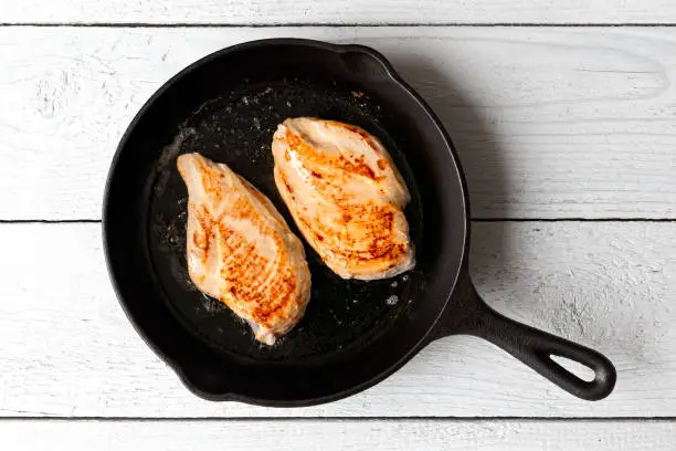 Two whole chicken breasts roasting in cast iron pan isolated on white wood. Top view.