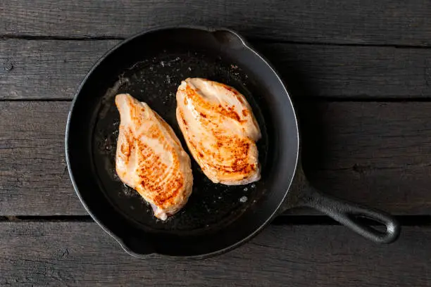 Two whole chicken breasts roasting in cast iron pan isolated on dark wood. Top view.