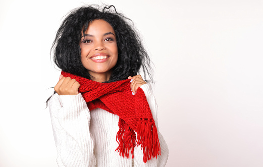 Close-up photo of cute African ethnic girl with messy dark hair, wearing a dazzling white sweater and red knitted scarf and holding it with her hands.
