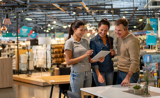 Cheerful sales woman showing a design on tablet to mid adult couple looking for furniture at a home store - Home renovation concepts