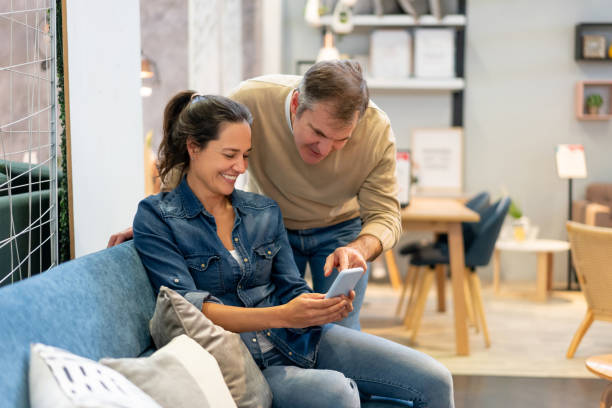 mid adult couple at a furniture store looking at something on smartphone talking and smiling - telephone indoors retail shopping mall imagens e fotografias de stock