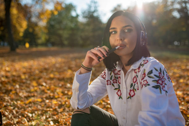 Young woman sitting in the park and smoking electronic cigarette stock photo