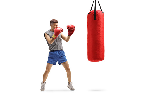 Full length shot of a young guy punching with boxing gloves in a red hanging bag isolated on white background