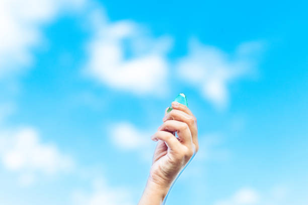 Vibrant green sea glass is being held by a child’s hand up to the clear blue sky filled with white fluffy clouds. A treasure found at the beach being held aloft. An image of hope and luck