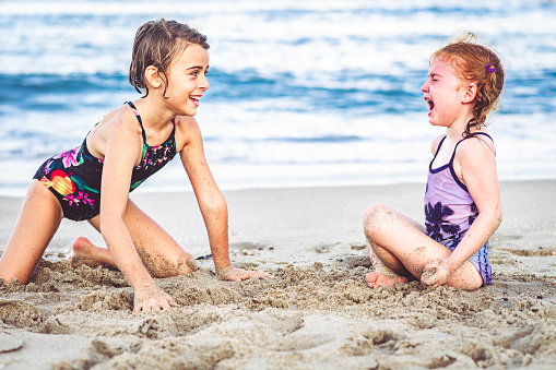 Adorable girls, sisters, play together in the sand at an idyllic beach. One is upset and crying, something went wrong and she is not pleased. Kids fighting, sibling rivalry or a broken friendship maybe. They are wearing swimwear maybe on a family vacation